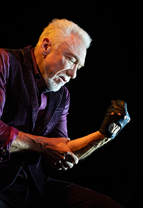 Patrick Page in All the Devils Are Here 6 Patrick Page wearing a purple shirt and black vest, sitting and leaning forward looks at the black leather fingerless glove on his clinched left hand as his right hand grips the left arm's elbo